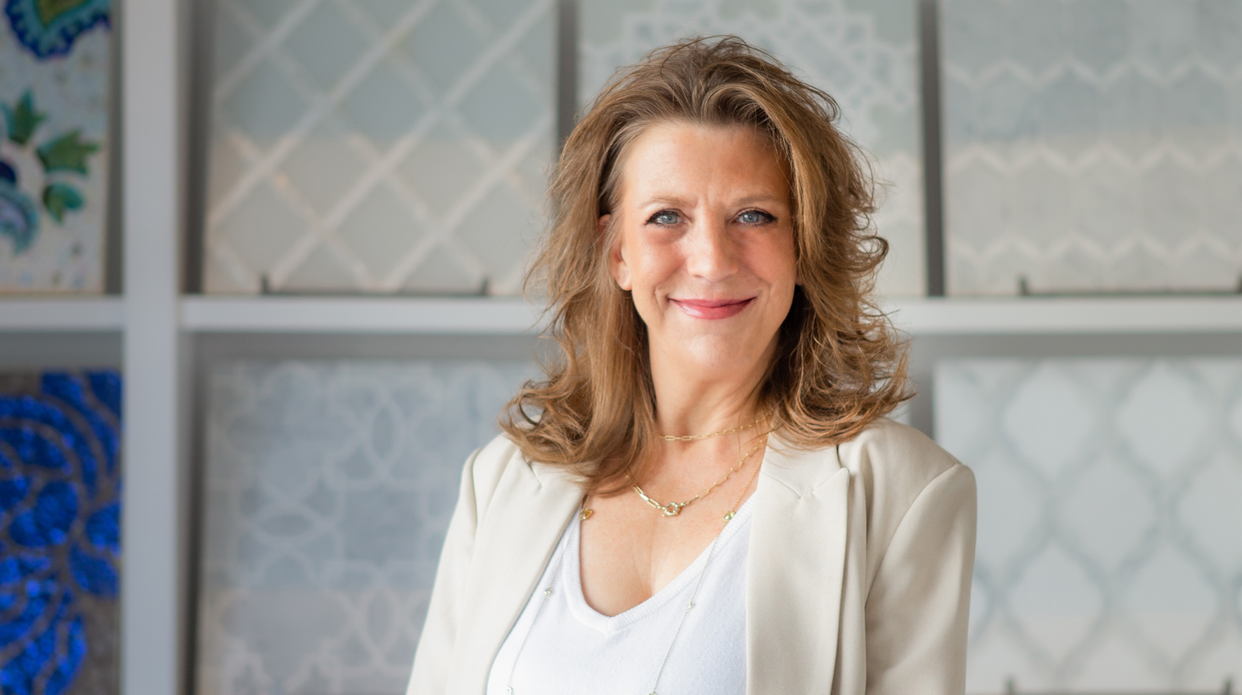 A portrait of a smiling woman with wavy hair, wearing a white top and beige blazer, standing in front of decorative wall art.