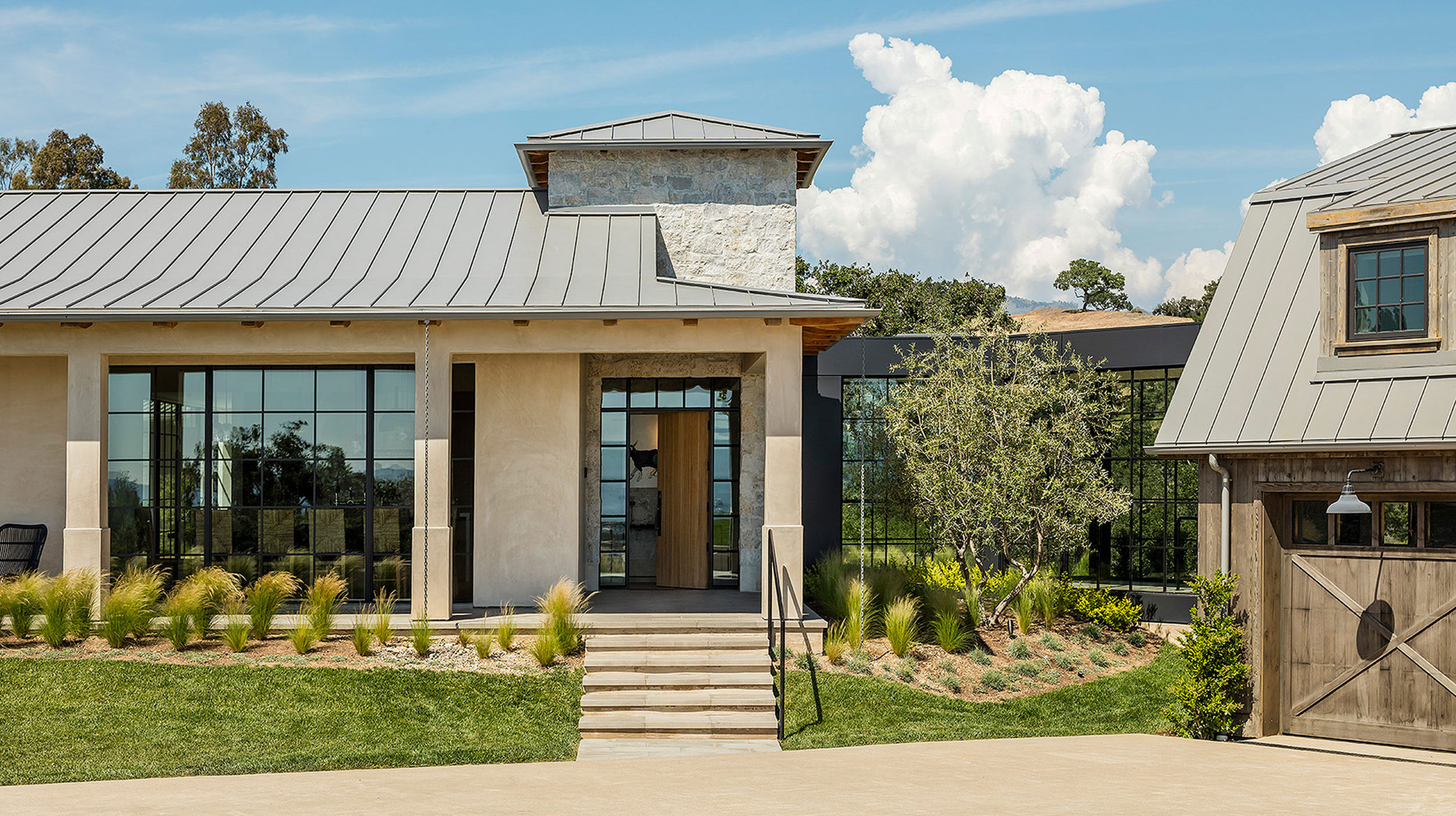 A modern farmhouse exterior with metal roofing, large windows, stone and stucco walls, landscaped garden, and a wooden garage door.