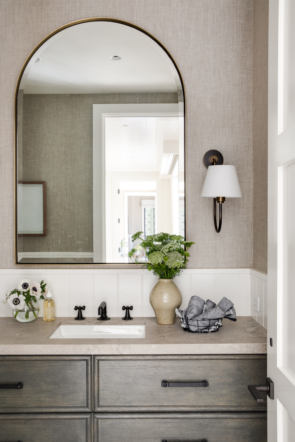 A stylish bathroom with a large arched mirror, black fixtures, a beige countertop, wooden vanity, sconce lighting, and decorative flowers.