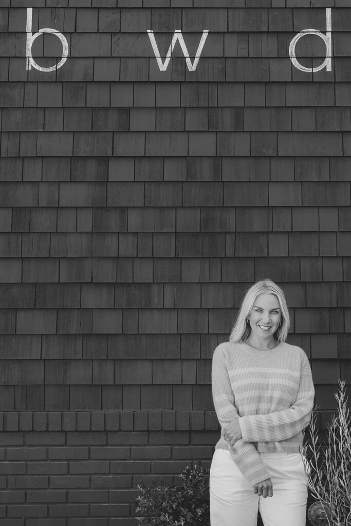 A black-and-white photo of a smiling woman standing in front of a dark shingle wall with the letters "b w d" written above her.