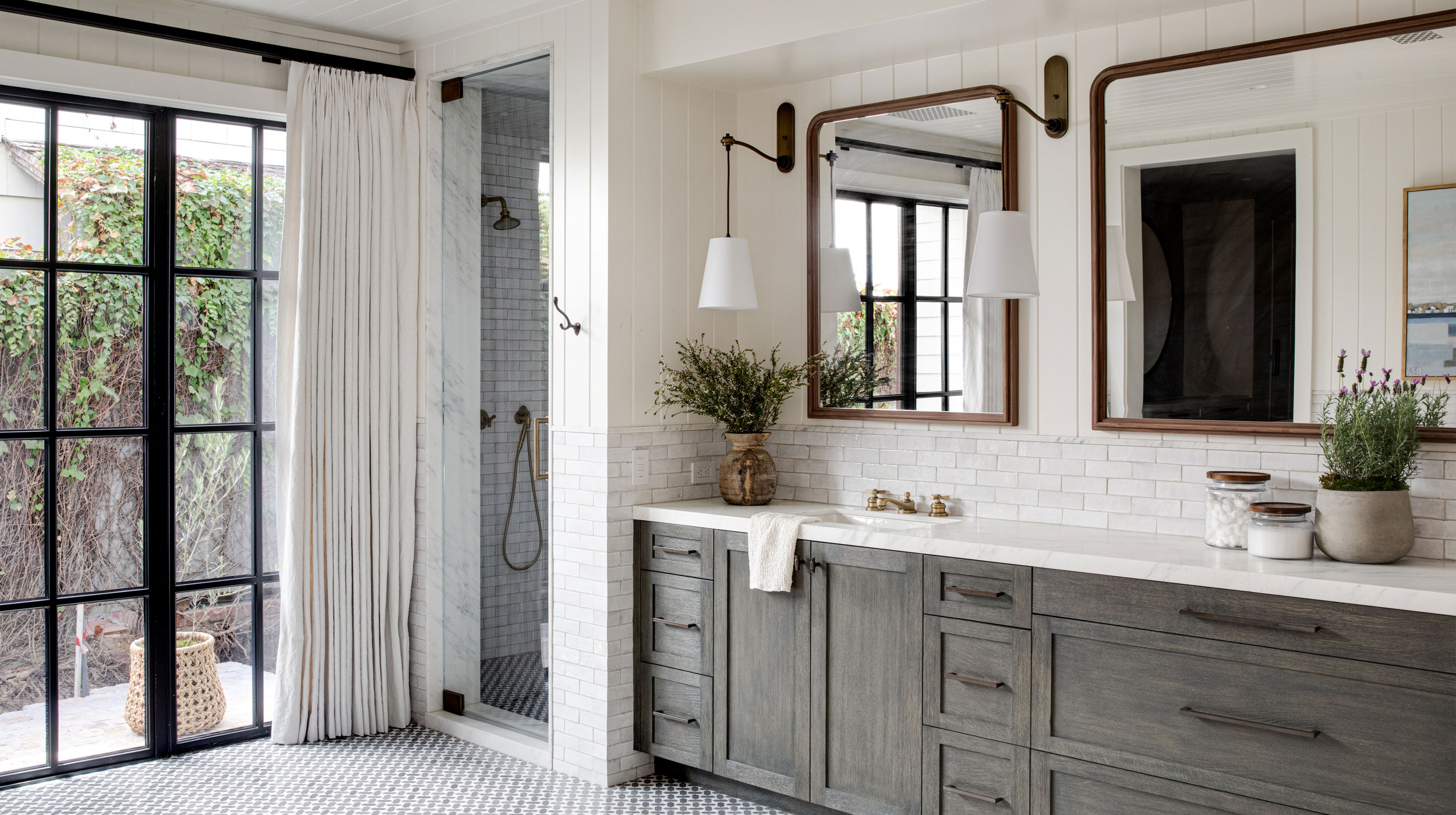 A bright bathroom with a large vanity, gray cabinets, dual mirrors, a walk-in shower, black-framed glass doors, and white curtains.