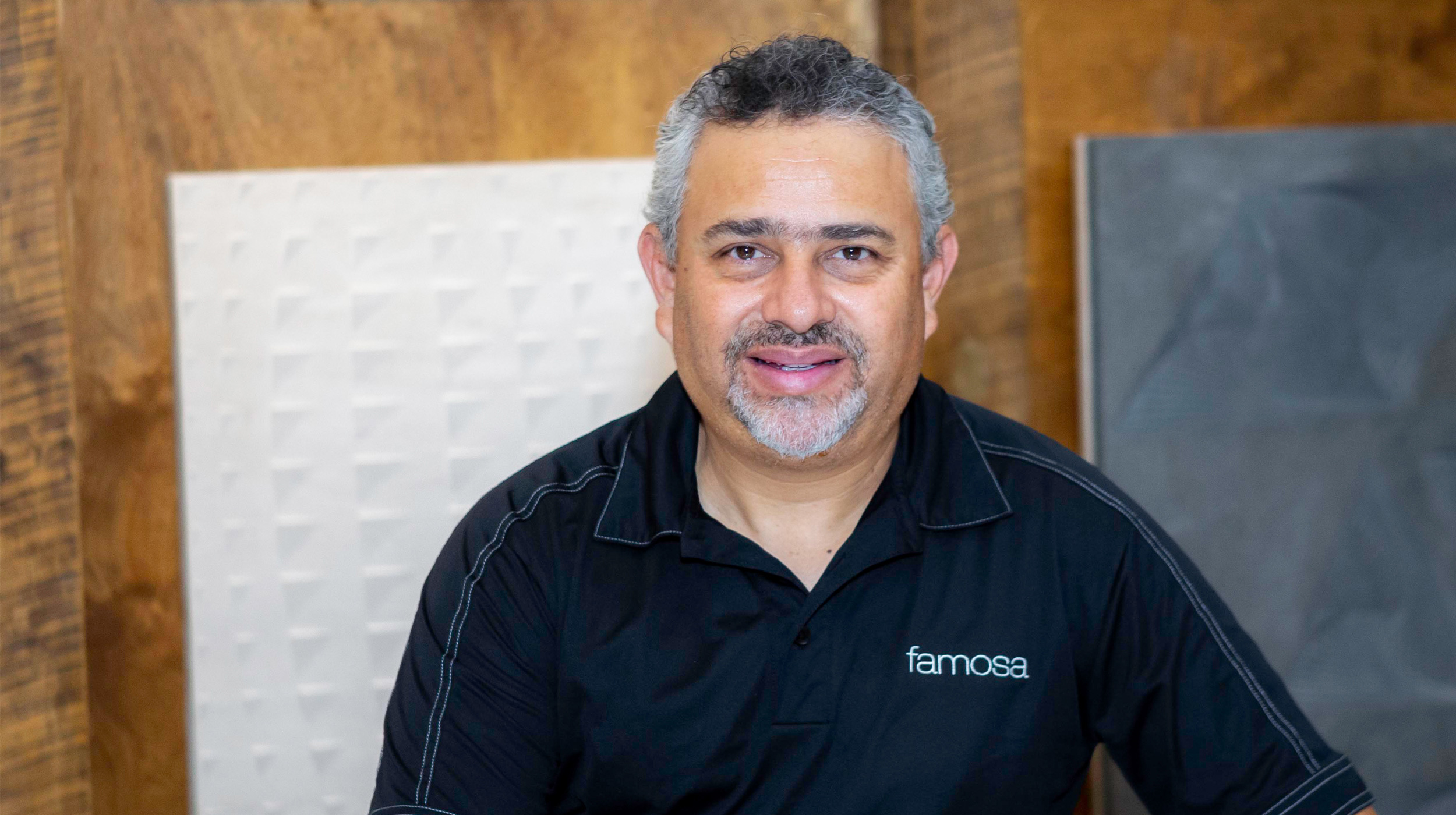 A portrait of a man with short, curly hair and a beard, wearing a black shirt with "famosa" logo, standing in front of textured wall panels.