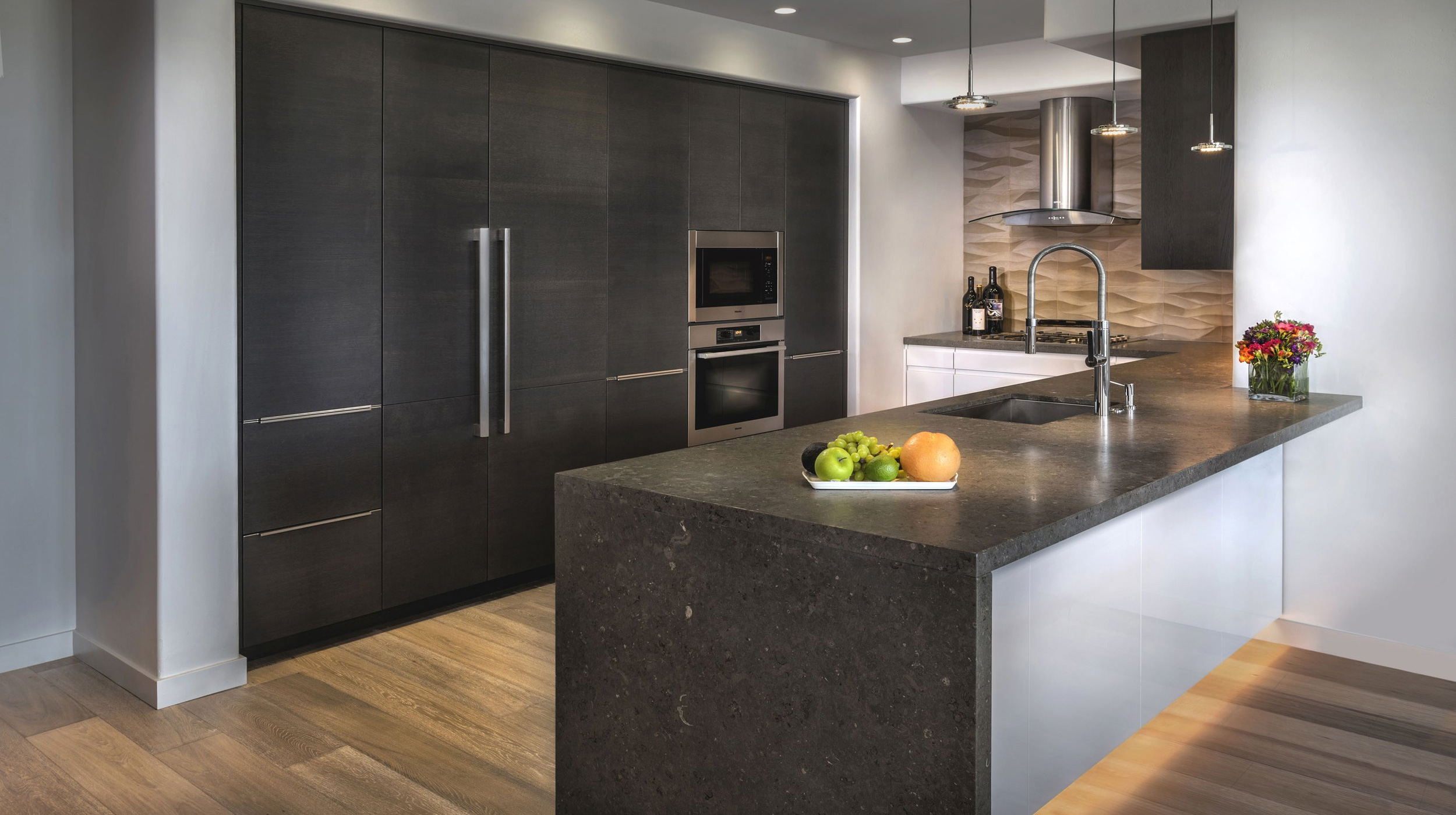 A sleek kitchen with dark cabinetry, stainless steel appliances, a large island with a dark countertop, modern pendant lights, and wooden flooring.