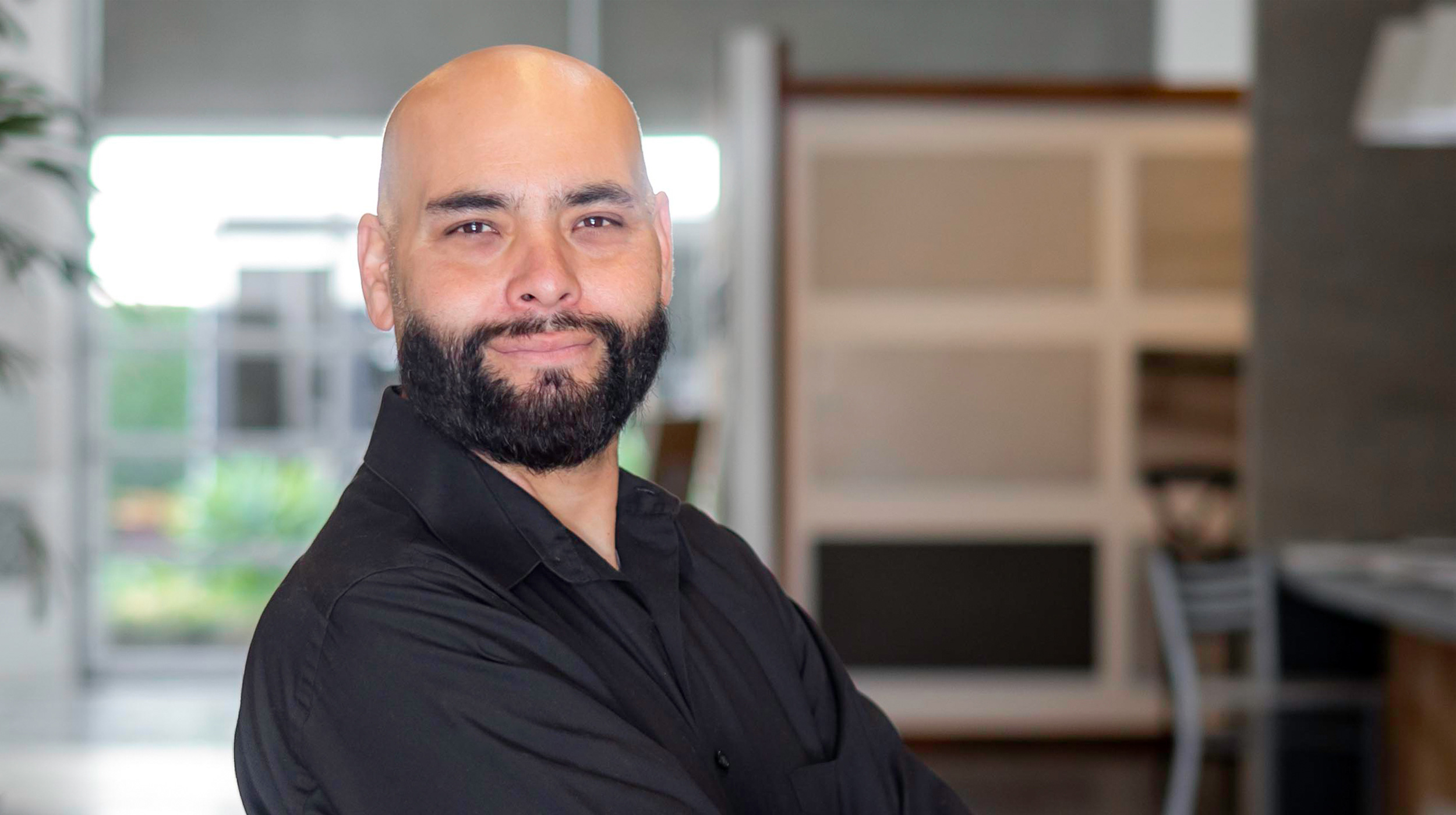 A portrait of a bearded man with a bald head, wearing a black shirt, smiling confidently with arms crossed, in a modern indoor setting.