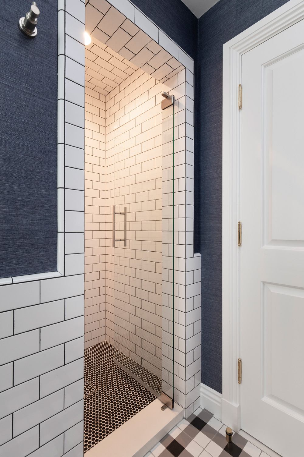 Modern shower with white subway tiles, black grout, a glass door, black penny tile floor, and adjacent white door with gold hinges.