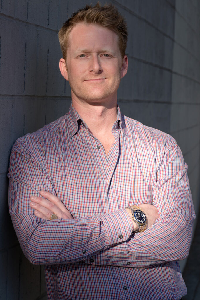 A man with light hair in a red and blue checkered shirt stands with his arms crossed against a textured wall, wearing a gold wristwatch and a confident expression.