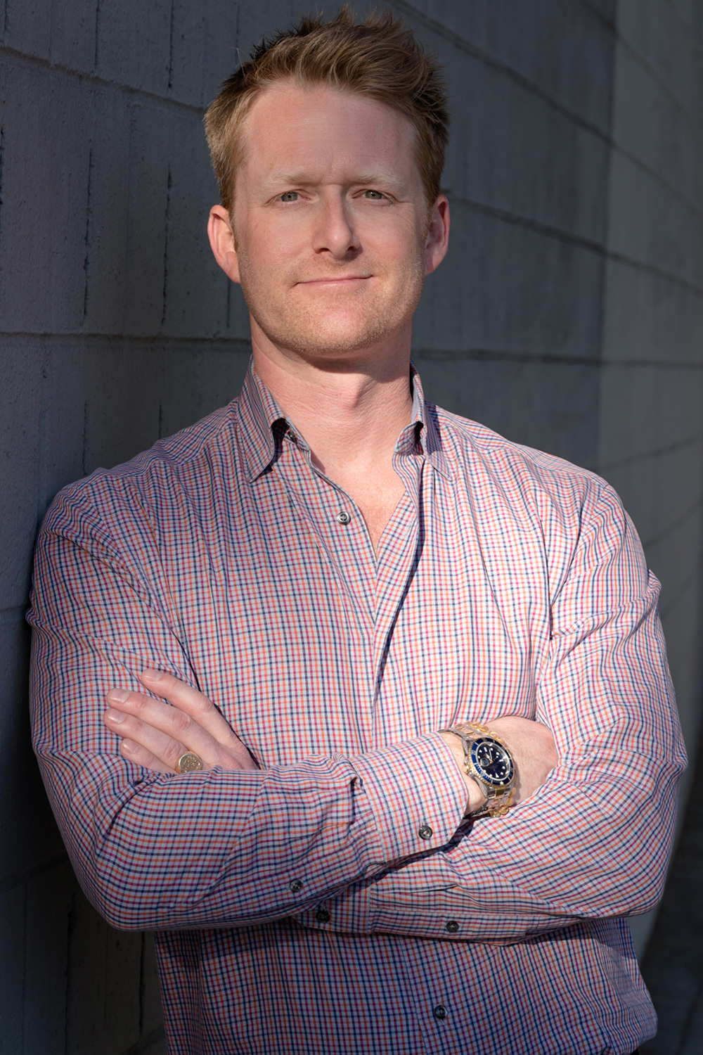 A man with light hair in a red and blue checkered shirt stands with his arms crossed against a textured wall, wearing a gold wristwatch and a confident expression.