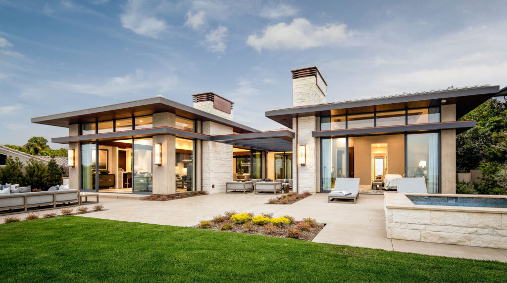 A modern home exterior featuring large windows, flat roofs, stone walls, manicured lawn, patio seating, and a small pool.