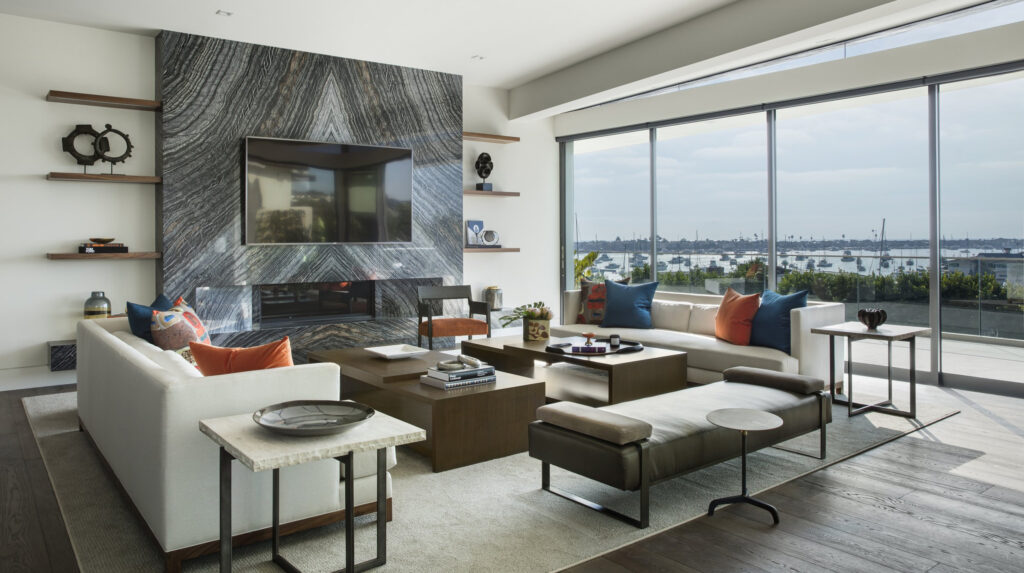 A modern living room with large windows offering a water view, featuring a marble accent wall with a TV, white sofas, colorful pillows, and wooden shelves.