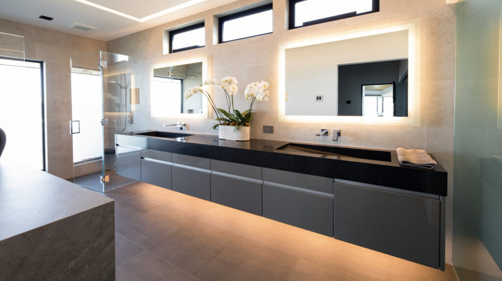 A contemporary bathroom with floating vanity, black countertop, backlit mirrors, glass shower, beige tiles, and an orchid arrangement.