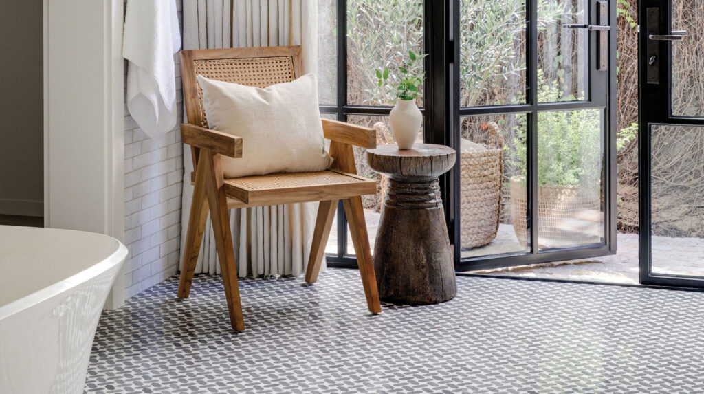 A serene bathroom corner with a wooden chair, a white pillow, a small rustic table, and black-framed glass doors opening to a garden.