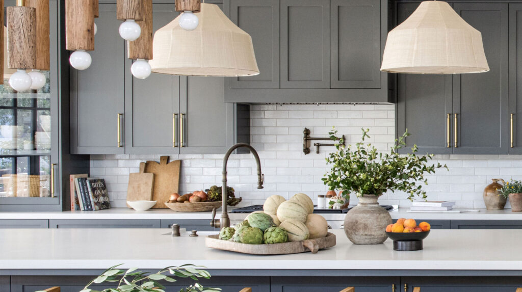 A modern kitchen with gray cabinets, white countertops, wooden and woven pendant lights, decorative plants, and a central island.