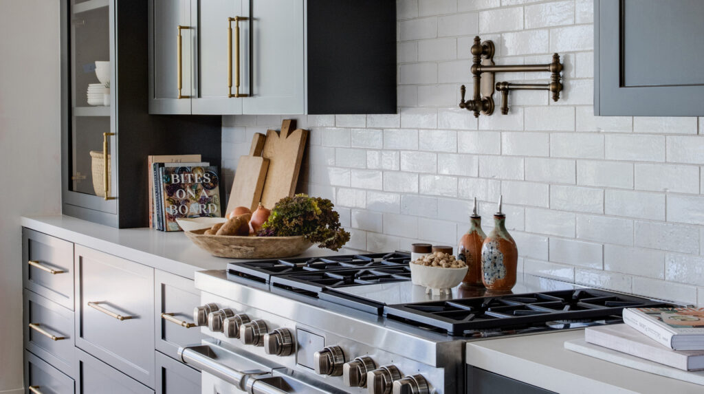 A modern kitchen with gray cabinets, brass hardware, a stainless steel stove, white subway tile backsplash, and decorative items.