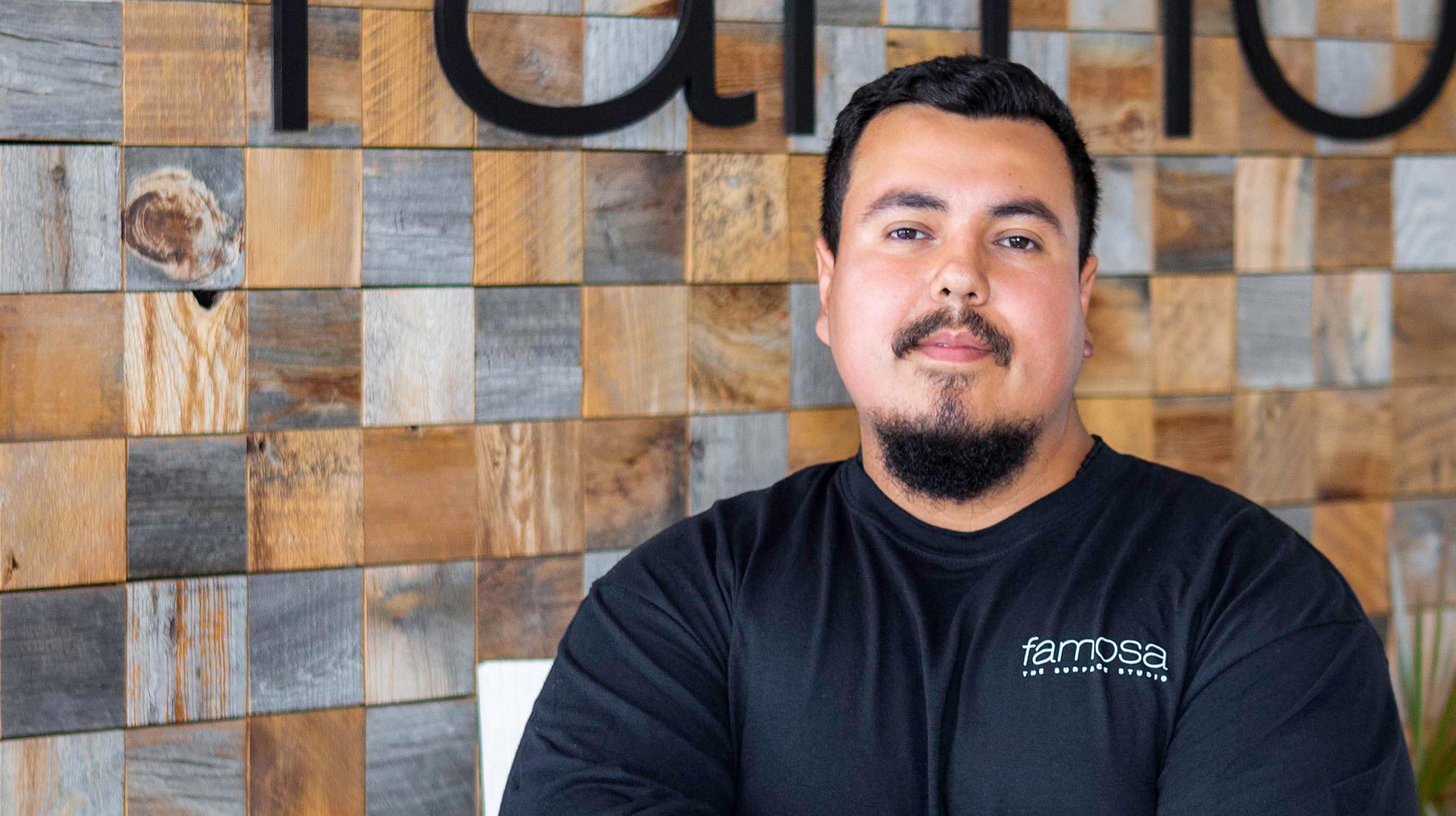 A portrait of a man with short hair and a beard, wearing a black "famosa" logo shirt, standing in front of a wooden mosaic wall with large black letters.