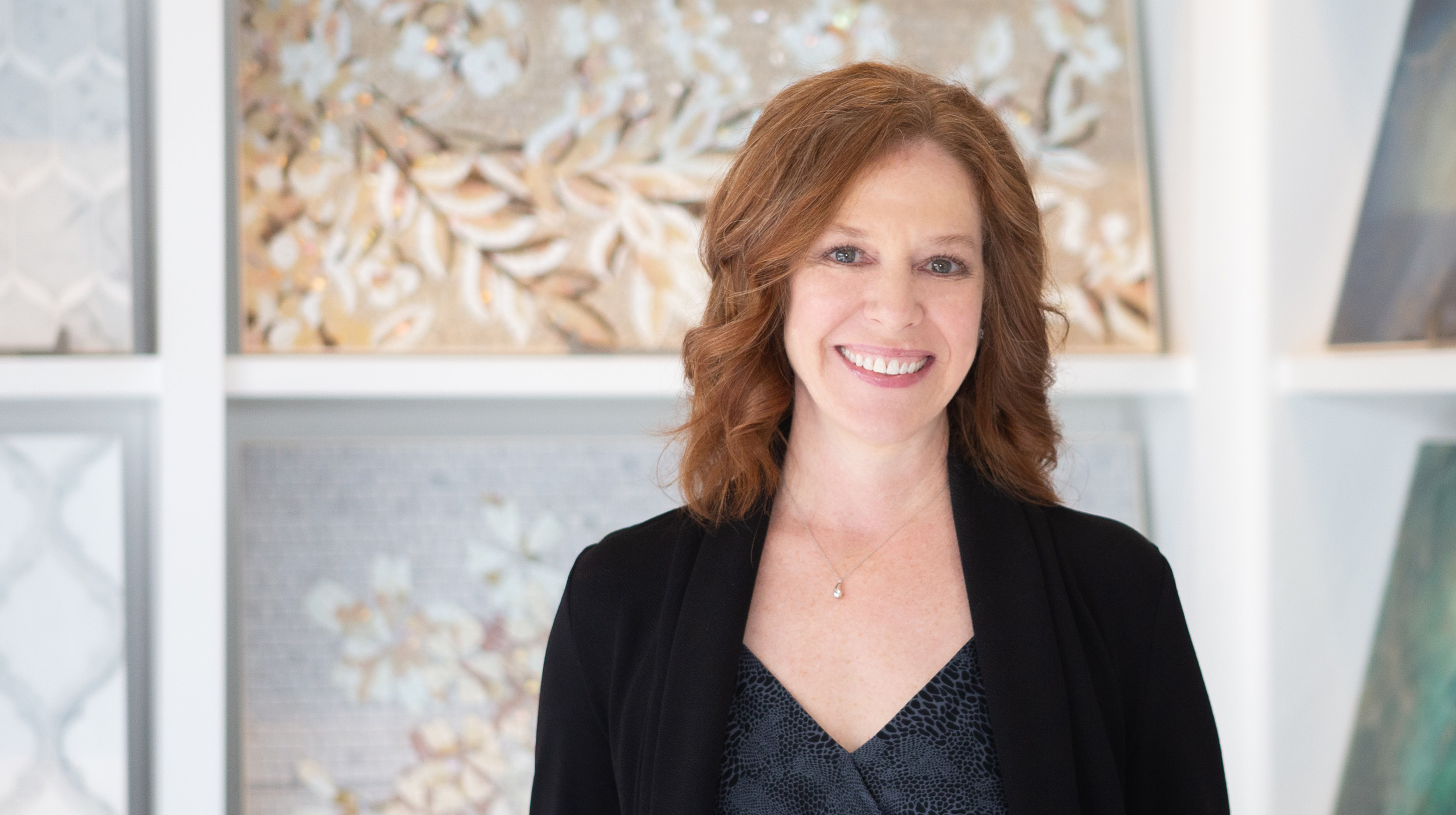 A portrait of a smiling woman with shoulder-length wavy hair, wearing a black top and necklace, standing in front of decorative wall art.