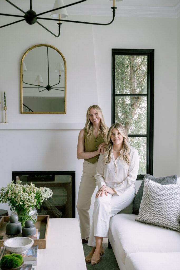 Two women in a stylish living room, one standing and one sitting on a white sofa, with a large mirror, modern decor, and large windows.