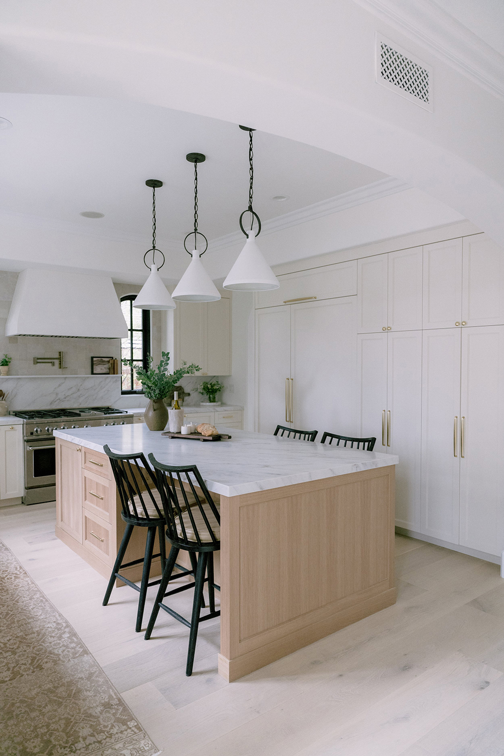 A modern kitchen with a large marble island, light wooden cabinetry, black chairs, and white pendant lights, creating a bright, airy space.