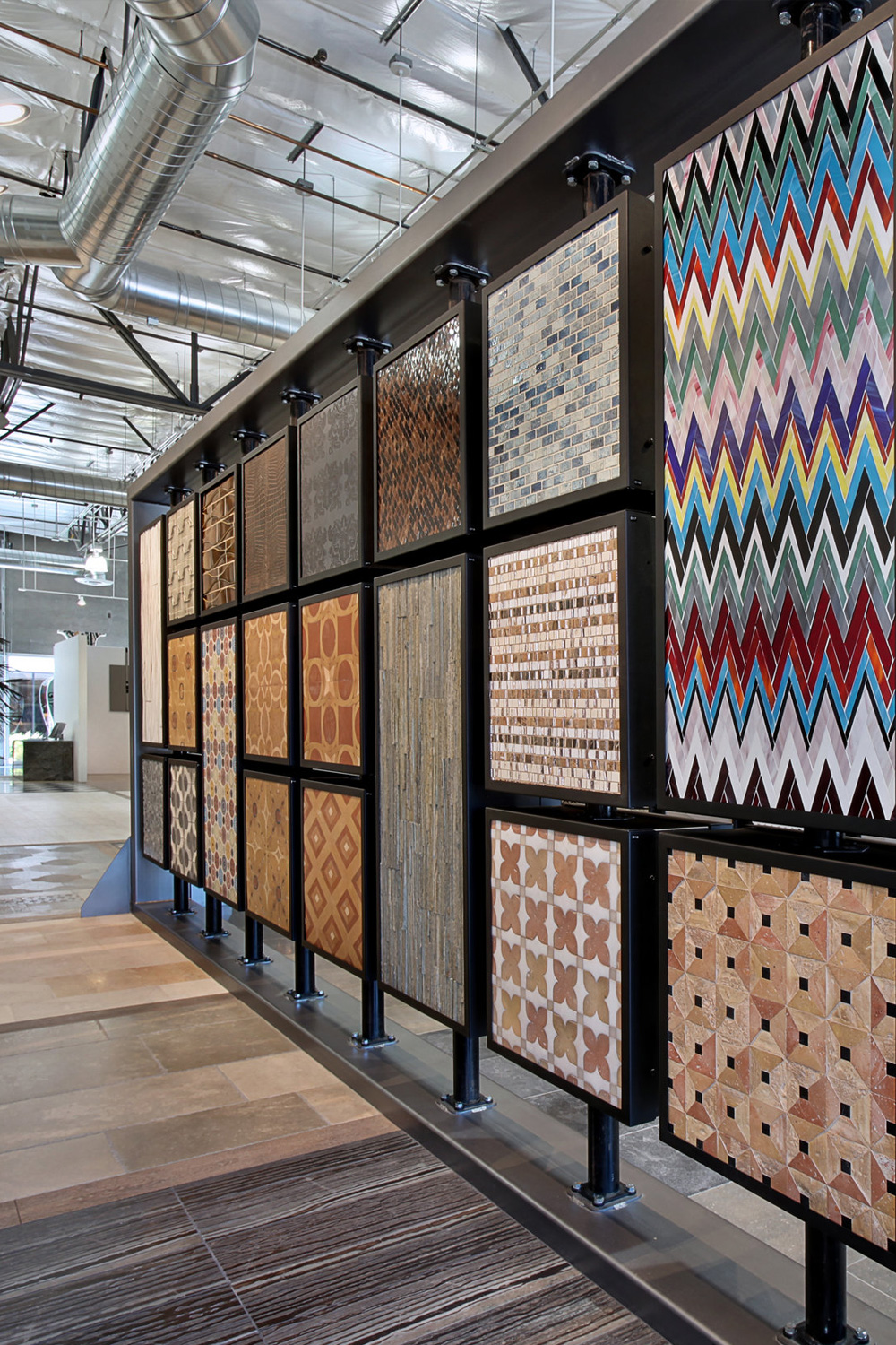 A display wall featuring various tile samples in a showroom, showcasing different patterns, textures, and colors under a high ceiling with industrial ductwork.