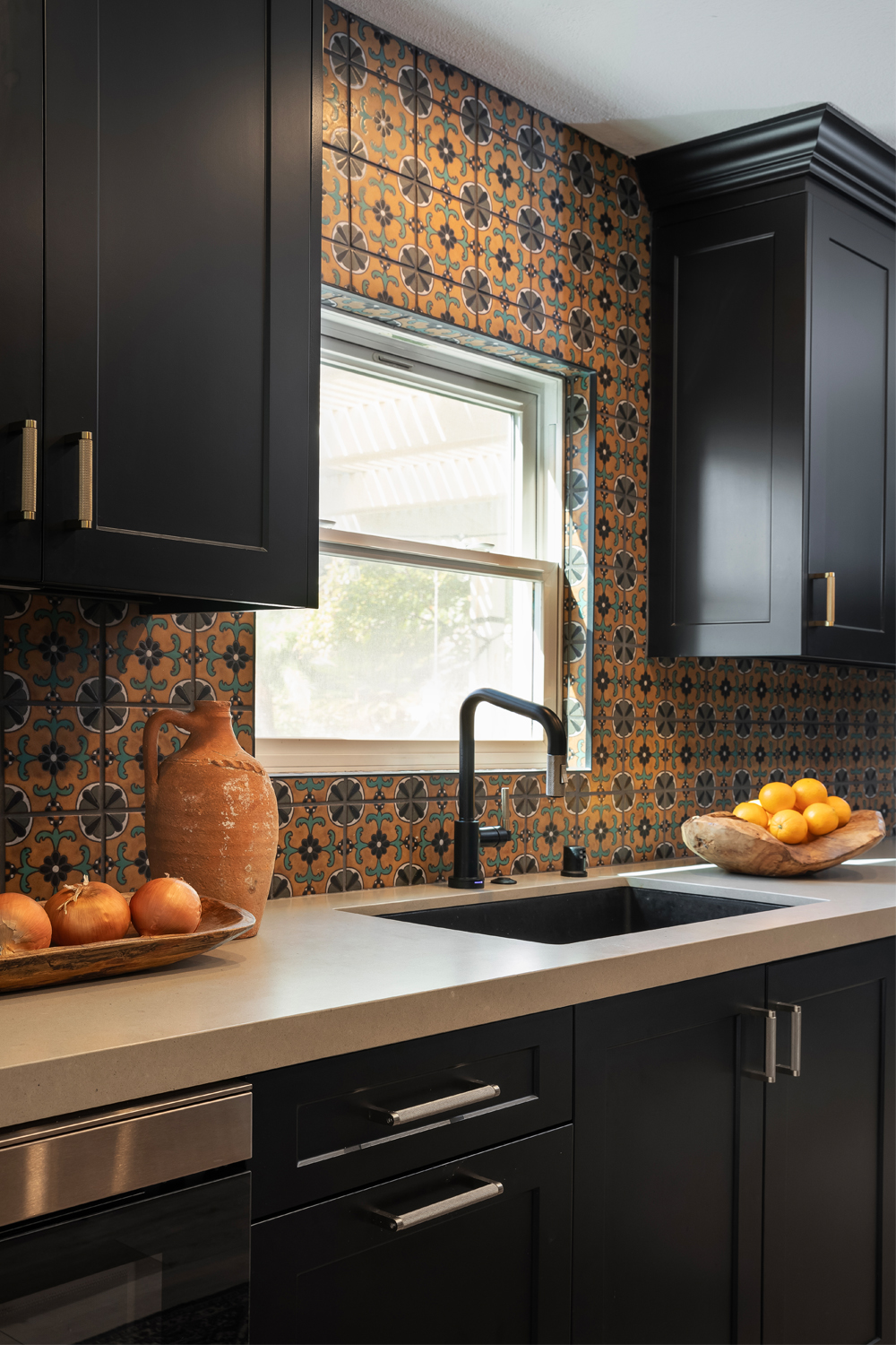 A kitchen with dark cabinets, a patterned tile backsplash, a black sink, and countertops with bowls of oranges and onions, under a window.