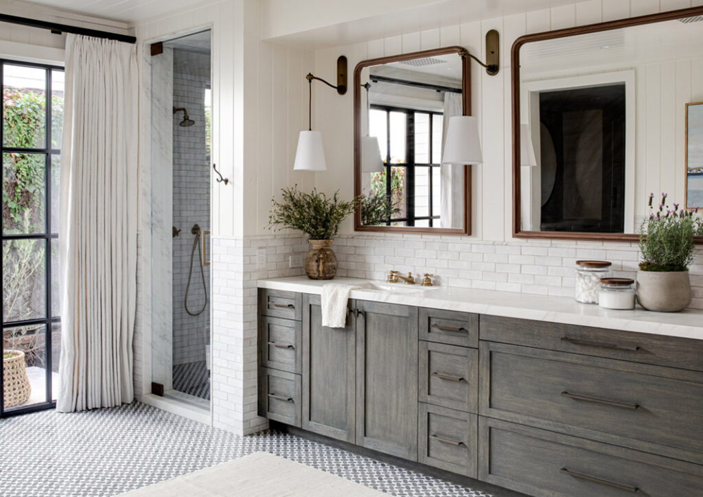A spacious bathroom with a large vanity featuring gray cabinets, white countertop, brass fixtures, dual mirrors, and a walk-in shower.