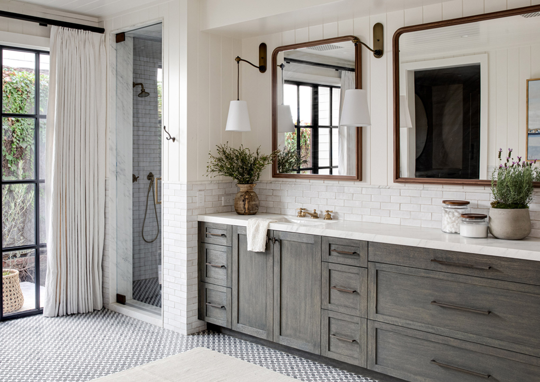 A spacious bathroom with a large vanity featuring gray cabinets, white countertop, brass fixtures, dual mirrors, and a walk-in shower.