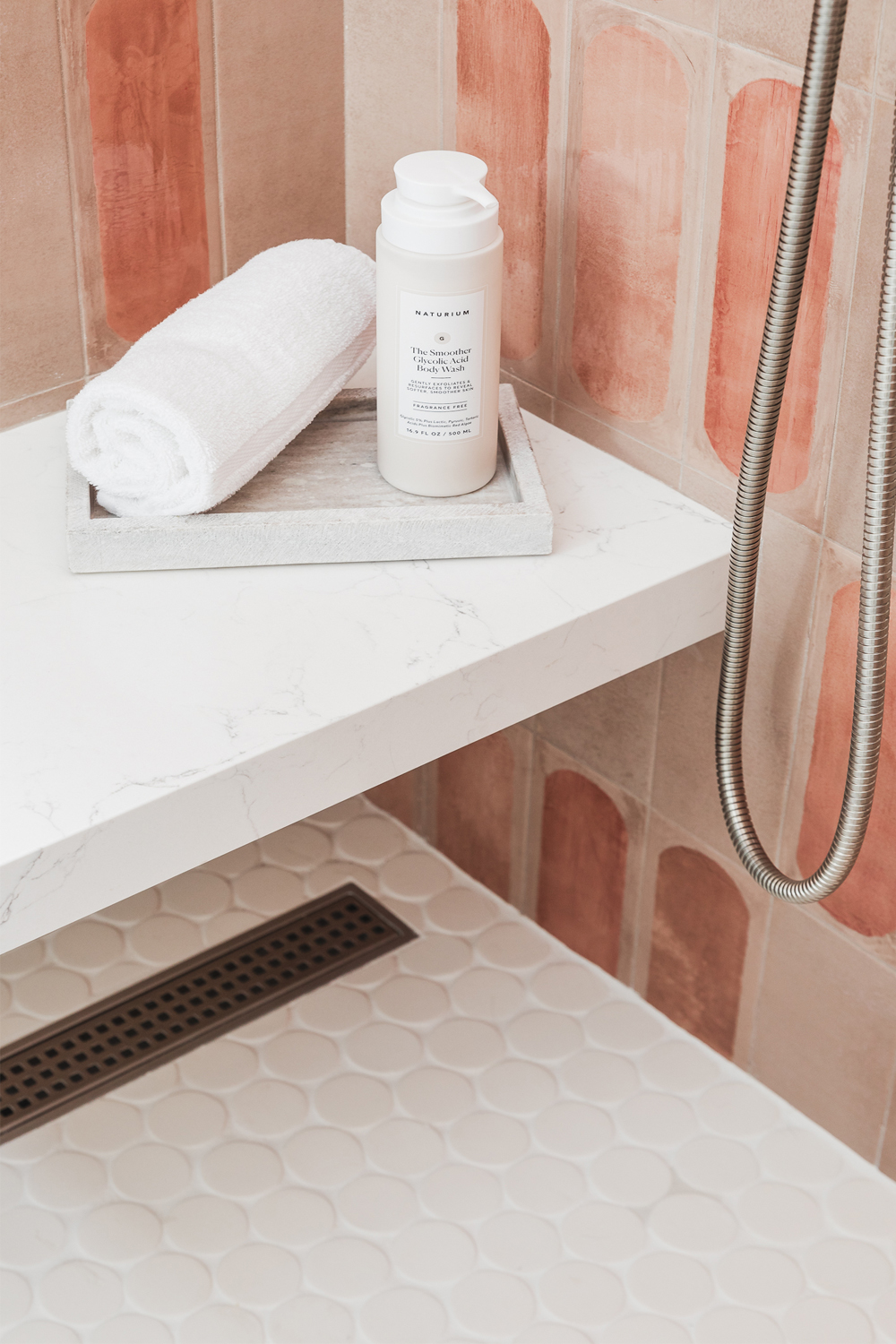 A shower corner with white penny round floor tiles, a marble shelf holding a rolled towel and body wash, and orange accent wall tiles.